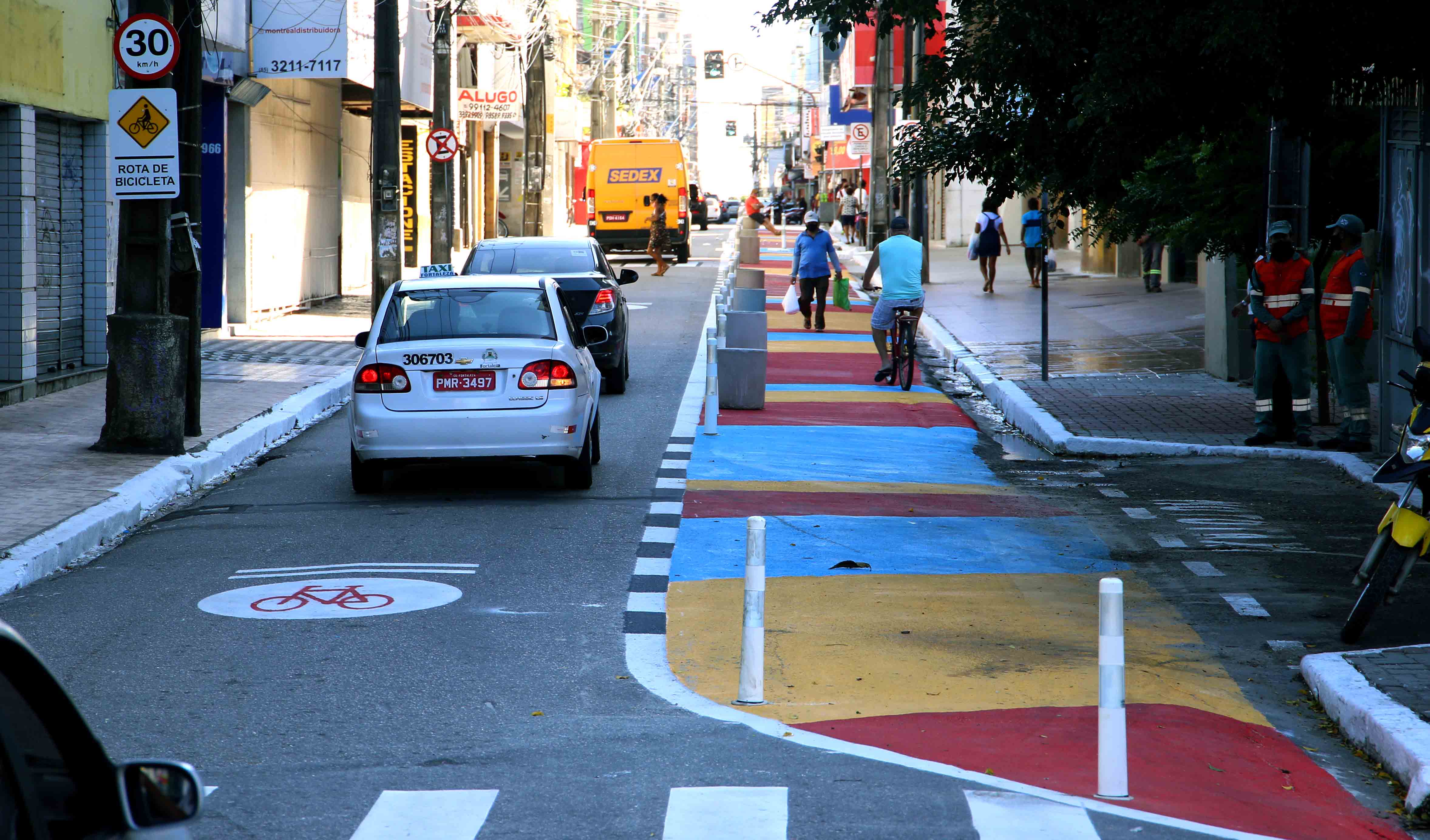 a foto mostra uma rua do centro do carros e pedestres, o detalhe é que parte da via está pintada de amarelo e vermelho e com cones para que as pessoas possam circular em segurança